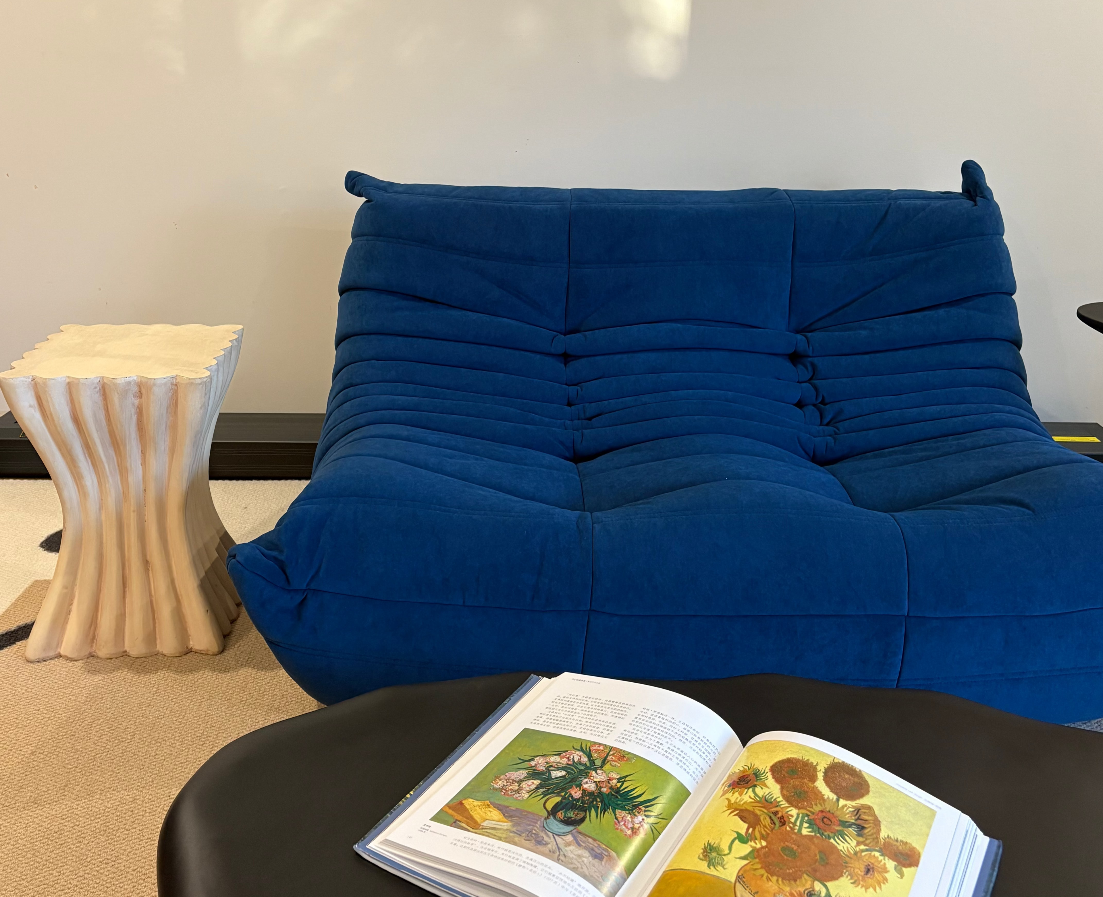 Blue sofa with an open book featuring Van Gogh's 'Sunflowers' on a black table, against a white wall with a painting.