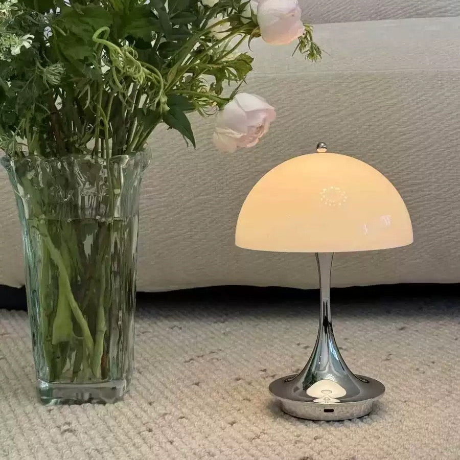 A mushroom-shaped table lamp with a glowing warm white dome shade and a polished silver metal base, placed on a textured beige carpet next to a glass vase with flowers in front of a light-colored sofa.