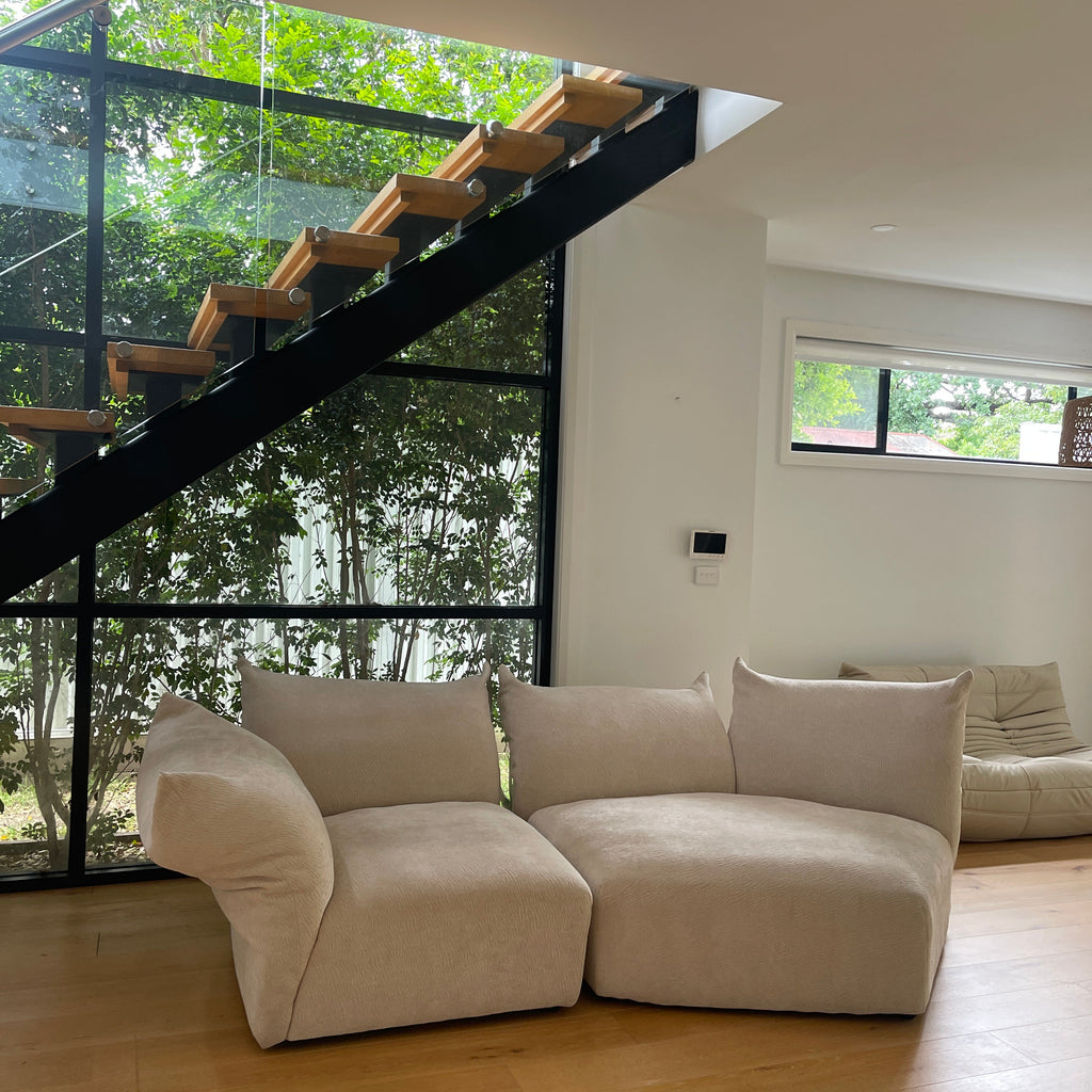Modern living room with a beige sectional sofa and a glass wall showing greenery outside.
