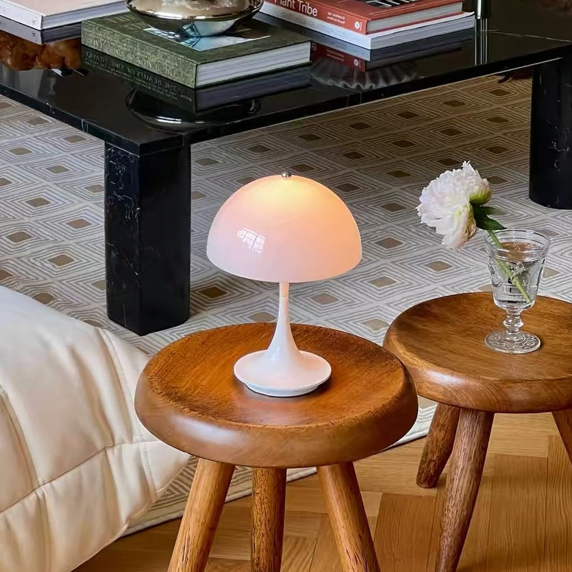 A styled living room scene featuring two round wooden side stools with tapered legs, one holding a white mushroom-shaped table lamp and the other holding a small glass vase with a white flower. In the background, a black marble-top coffee table holds books and a vase of flowers, set on a patterned rug over light wood flooring.