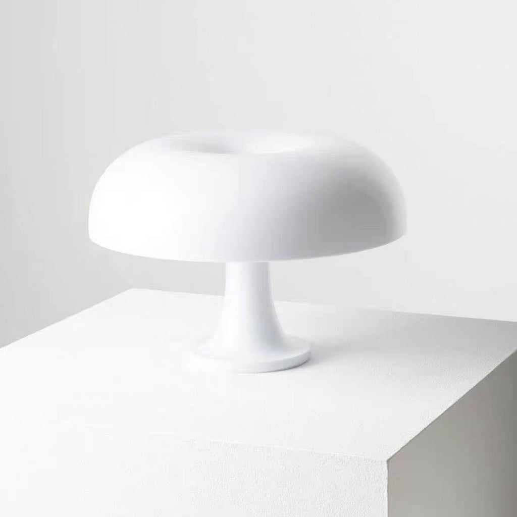 A minimalist white table lamp with a rounded, mushroom-like shade and a single pedestal base, displayed on a white cube pedestal against a light gray background.