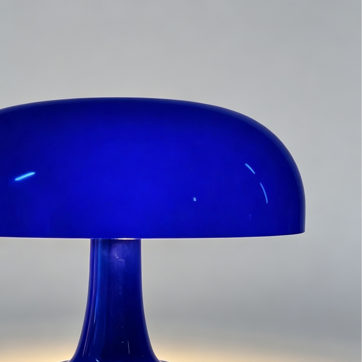 A glossy cobalt blue mushroom-shaped table lamp with a wide domed shade and a flared base, shown on a neutral gray background with warm light glowing from the base.