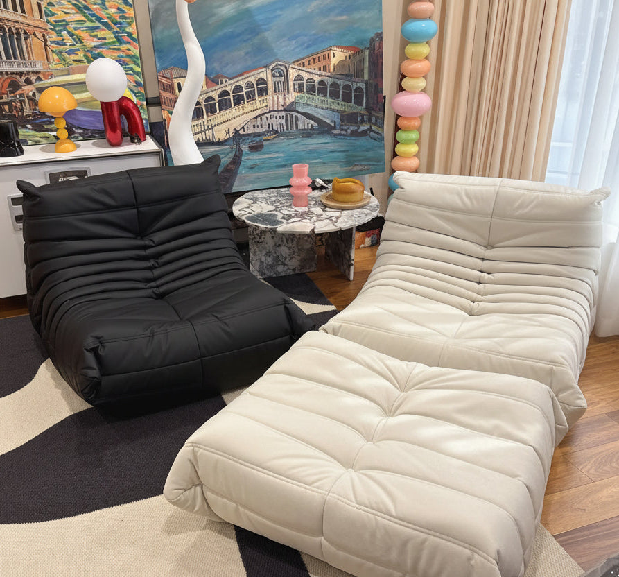 Beige lounge chair in a room with a black chair, colorful art, and a painting of a bridge.