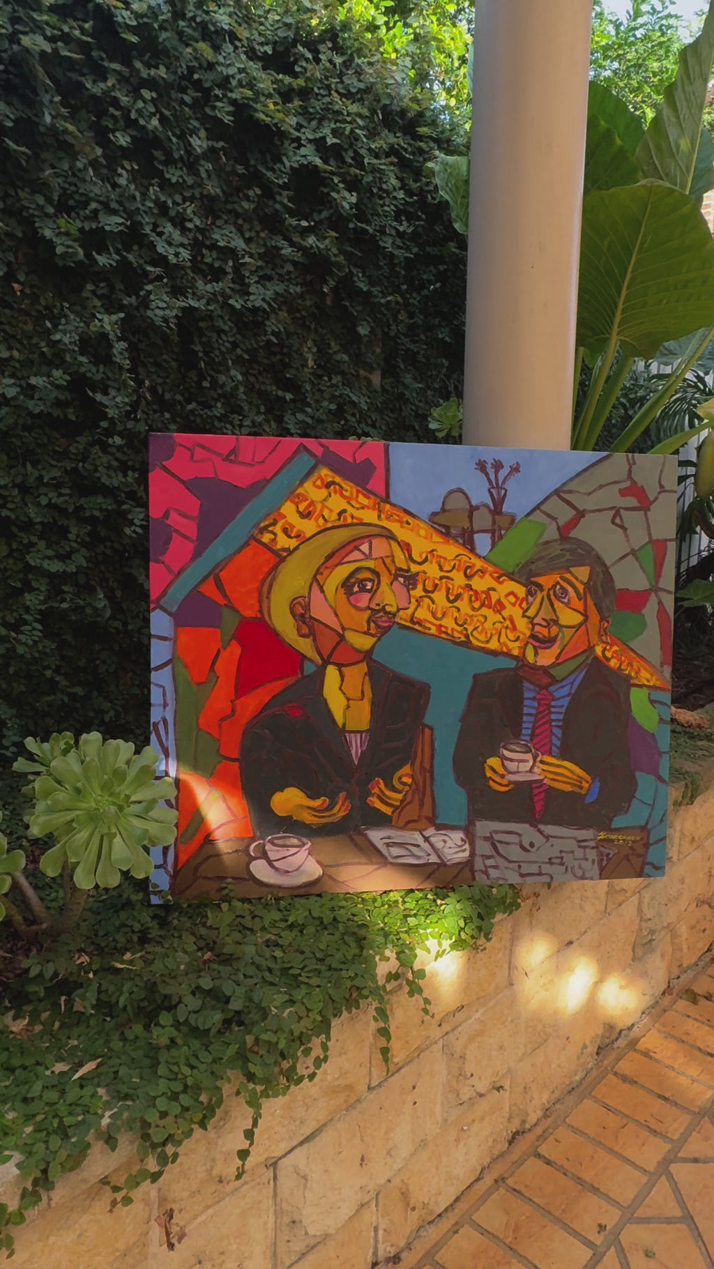 A bold and vibrant cubist painting featuring two figures engaged in conversation over coffee at a café. The artwork is rich with geometric shapes, expressive faces, and a striking colour palette of reds, yellows, and blues, evoking a lively and intellectual atmosphere.